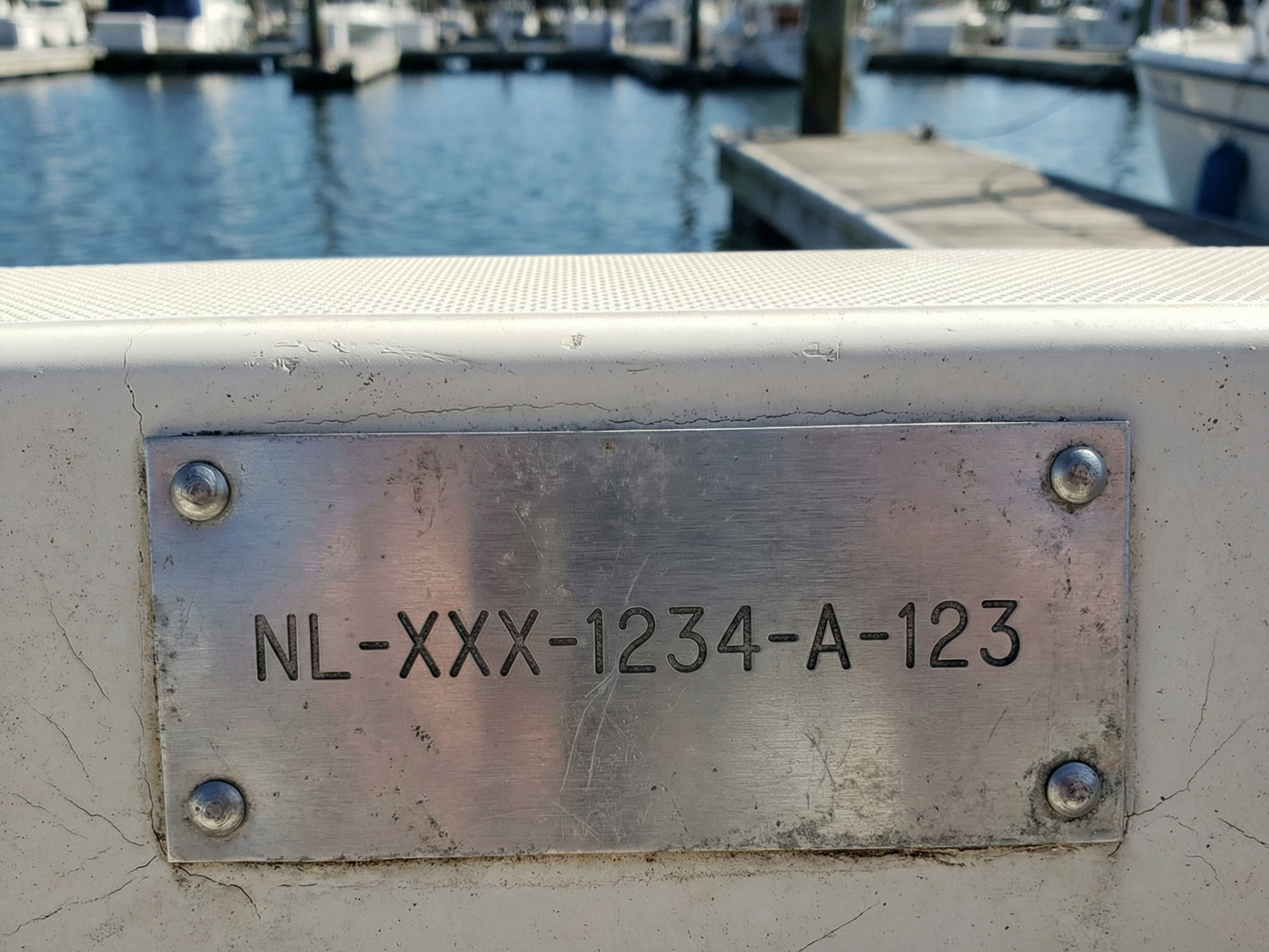 Een metalen plaat met het WIN-nummer 'NL-XXX-1234-A-123' bevestigd aan de reling van de spiegel van een boot. Op de achtergrond is een jachthaven met boten te zien onder een heldere hemel. Het WIN-nummer dient als uniek identificatienummer van de boot, met landcode, fabrikant, serienummer en bouwjaar.
